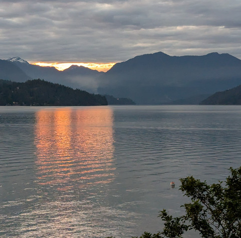 In the lower right, a few tree branches are visible and a buoy floating in the open water. Across that water, misty mountains and a turbulent cloudy sky. Between the mountains and the clouds, brilliant setting-sun light is visible and is reflected in the open water. 