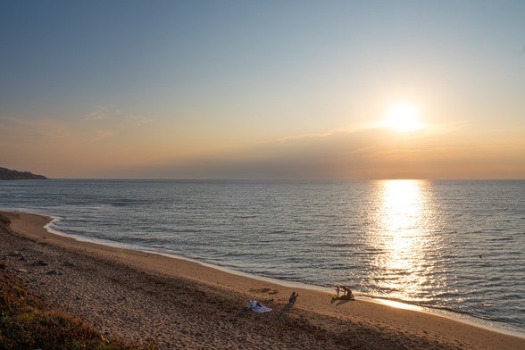 The sun is setting and, in the distance, a woman is taking a picture of a man and a child building a sandcastle on the beach.