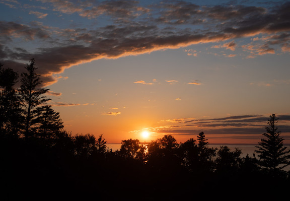 Sunset over the ocean, with a screen of trees below and an arc of clouds above.