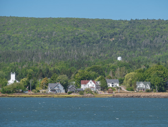 Looking across the ocean at a shoreline with houses along the waterfront, rising wooded hills behind, blue sky above.