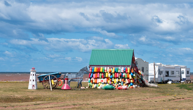 On the oceanfront, a small shed is covered with many brightly colored boat fenders. Also in the picture: Some vacation trailers, a statue of a horse and carriage, and a small front-lawn lighthouse decoration.