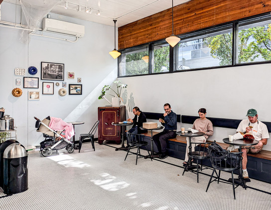 In a white cafe with wood trim, four individuals sit at adjacent tables, each back to the wall. An old bank safe is in the corner. Light comes  in the windows high on the wall behind the customers and casts shadows on the floor. Another wall has a pattern of hanging artworks; below them, a baby carriage with a pink cloth which presumably covers the baby.