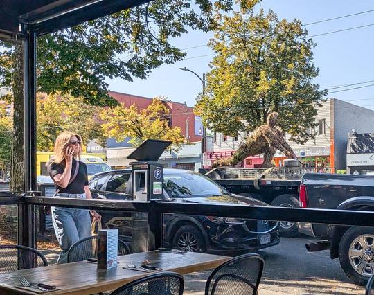From inside a cafe, a busy street. A woman in jeans and a black T-shirt is walking and talking on her mobile. On the other side of the dense traffic, store fronts and trees.  Looming over the cars - maybe on the back of a truck? - is a large vaguely human figure kneeling as if to start a race. Its surface seems organic: wood or plants?