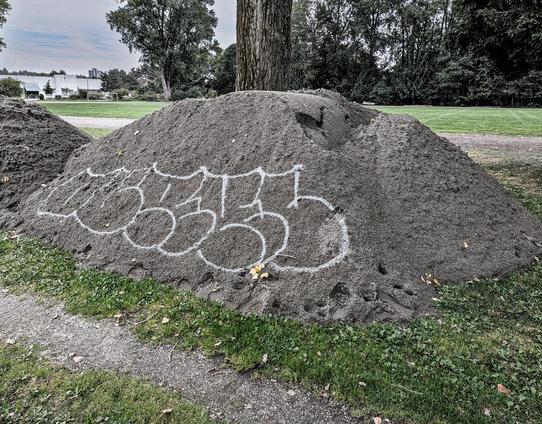 A heap of construction sand beside a narrow pathway, tree behind. On the sand heap, typically-illegible graffiti. 