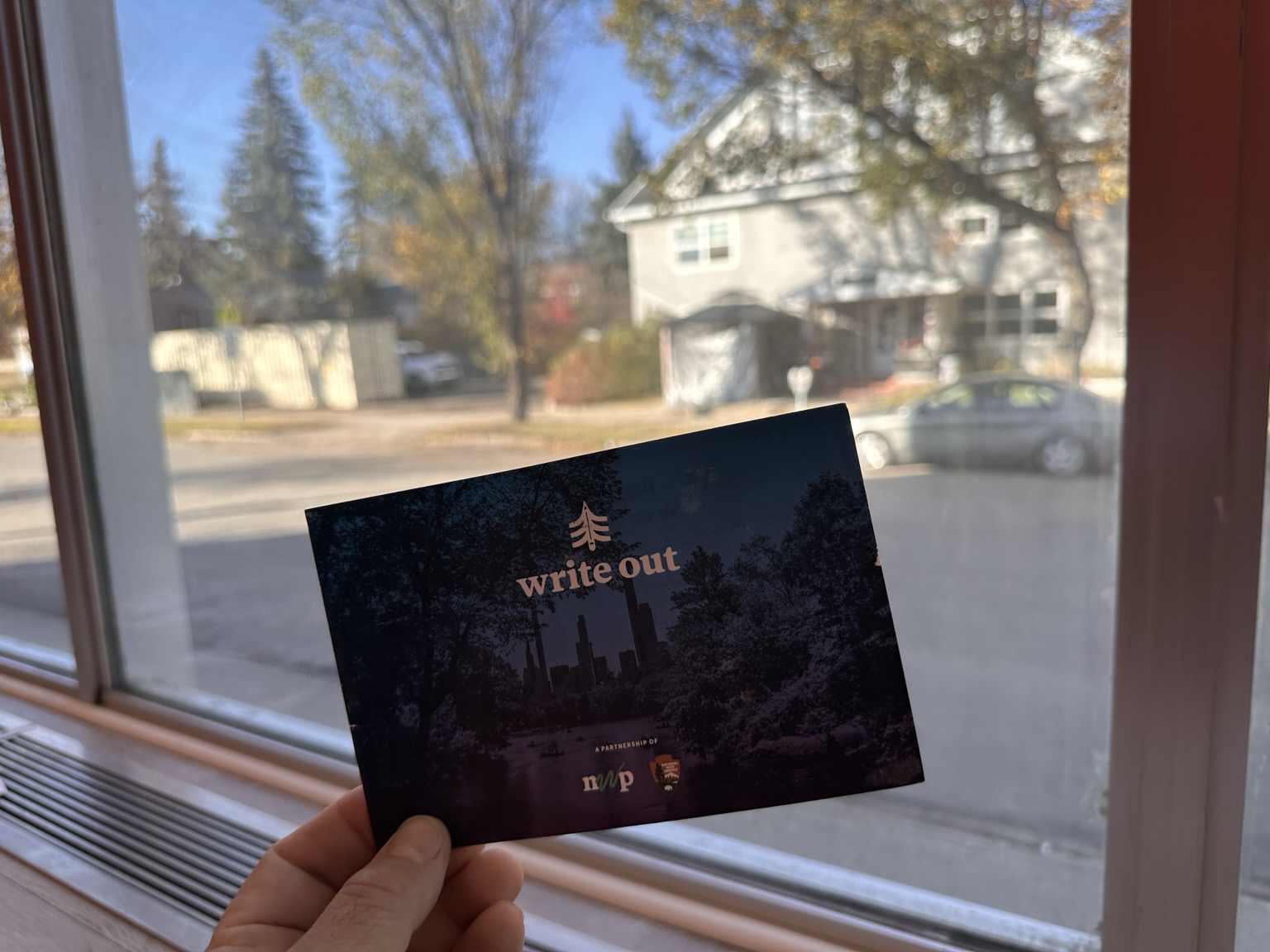 Hand holding a postcard bearing a write out text and logo, in front of window looking out at tree lined street and 3 story house.