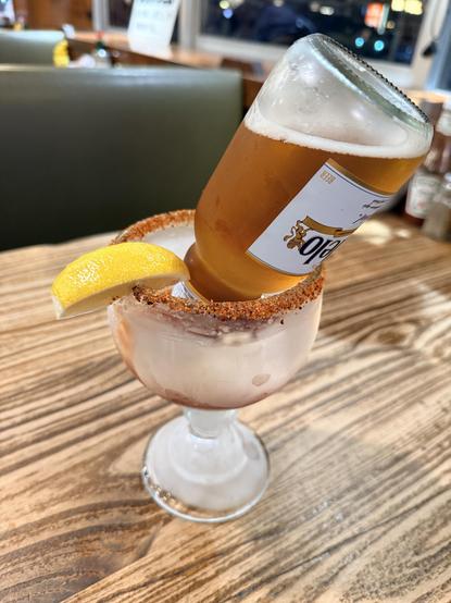 A wide frosted and spiced rim glass with an upside down bottle of modelo in a pool of tomato juice