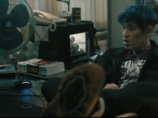 Screen Grab from Slow Hosrses season 2, Roddy Ho with shoes up on a cluttered disk featuring a large thick book entitled "XML".