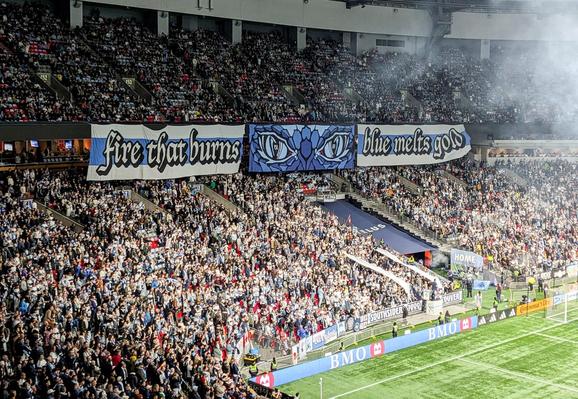 A portion of a large crowd at an indoor stadium, displaying a huge banner. Left of the banner is text “fire that burns”, center is a vaguely Tolkienesque photo of scary reptilian eyes, right is text "blue melts gold"